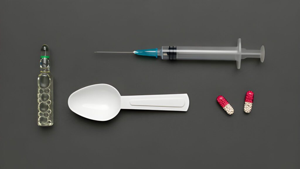 A syringe, ampoule, plastic spoon, and two red-and-white capsules on a dark gray background, conveying a medical or pharmaceutical theme.