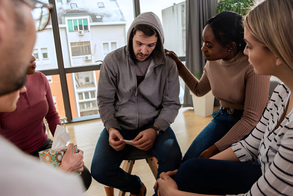 A group of people sit in a circle in a bright room. A man in a hoodie looks down, appearing distressed. Others offer support, one with a comforting hand on his shoulder.