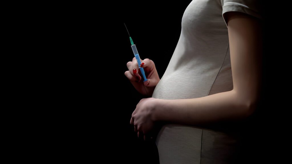 A pregnant woman holds a syringe in her hand, partially illuminated against a dark background. The atmosphere conveys caution and contemplation.