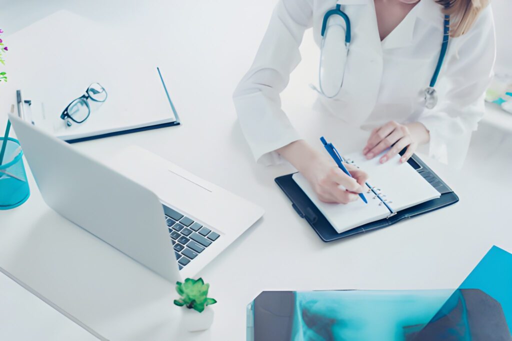 A doctor in a white coat, with a stethoscope, writes notes in a planner at a desk with a laptop, glasses, and an X-ray, conveying a focused, professional atmosphere.