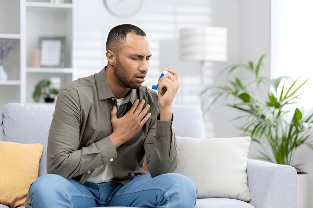 Man on a couch using a blue inhaler while clutching his chest, suggesting a respiratory problem or asthma attack.